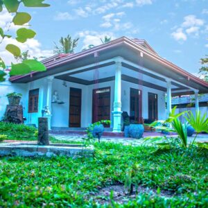 A single-storey house with a red tiled roof, surrounded by green grass and plants, under a bright blue sky.